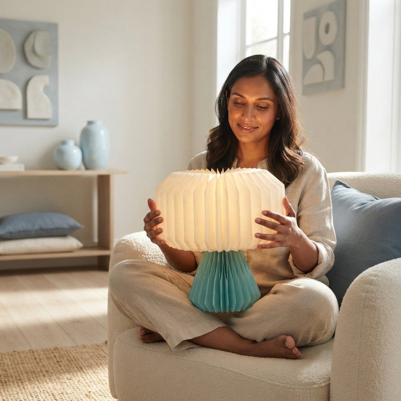 A South Asian woman holds an open, glowing, accordion-style paper lamp in a modern, sunlit living room with blue-toned decor.